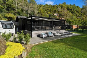 A modern outdoor seating area with a covered patio, a white caravan parked nearby, and a lush green landscape in the background. - Amalia Cottage - Lake Okareka, Huge Covered Deck, Sleeps 4 (Rotorua)