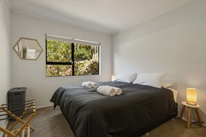 A bedroom with a large bed, a window with a view of greenery, a mirror, and a bedside lamp. - Amalia Cottage - Lake Okareka, Huge Covered Deck, Sleeps 4 (Rotorua)