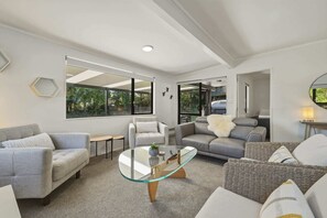 A modern living room with a glass coffee table, grey sofas, and a large window offering a view of greenery. - Amalia Cottage - Lake Okareka, Huge Covered Deck, Sleeps 4 (Rotorua)