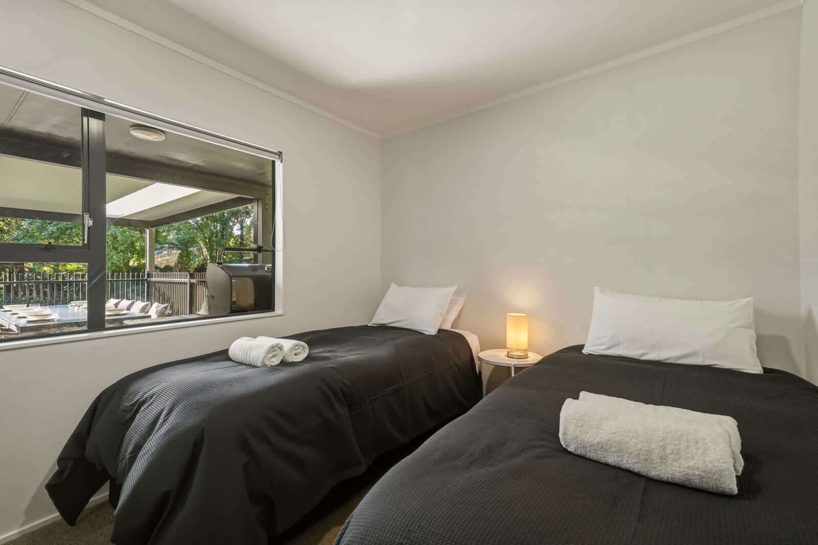 A hotel room with two beds, a window with a view of an outdoor seating area, and a small table with a lamp.