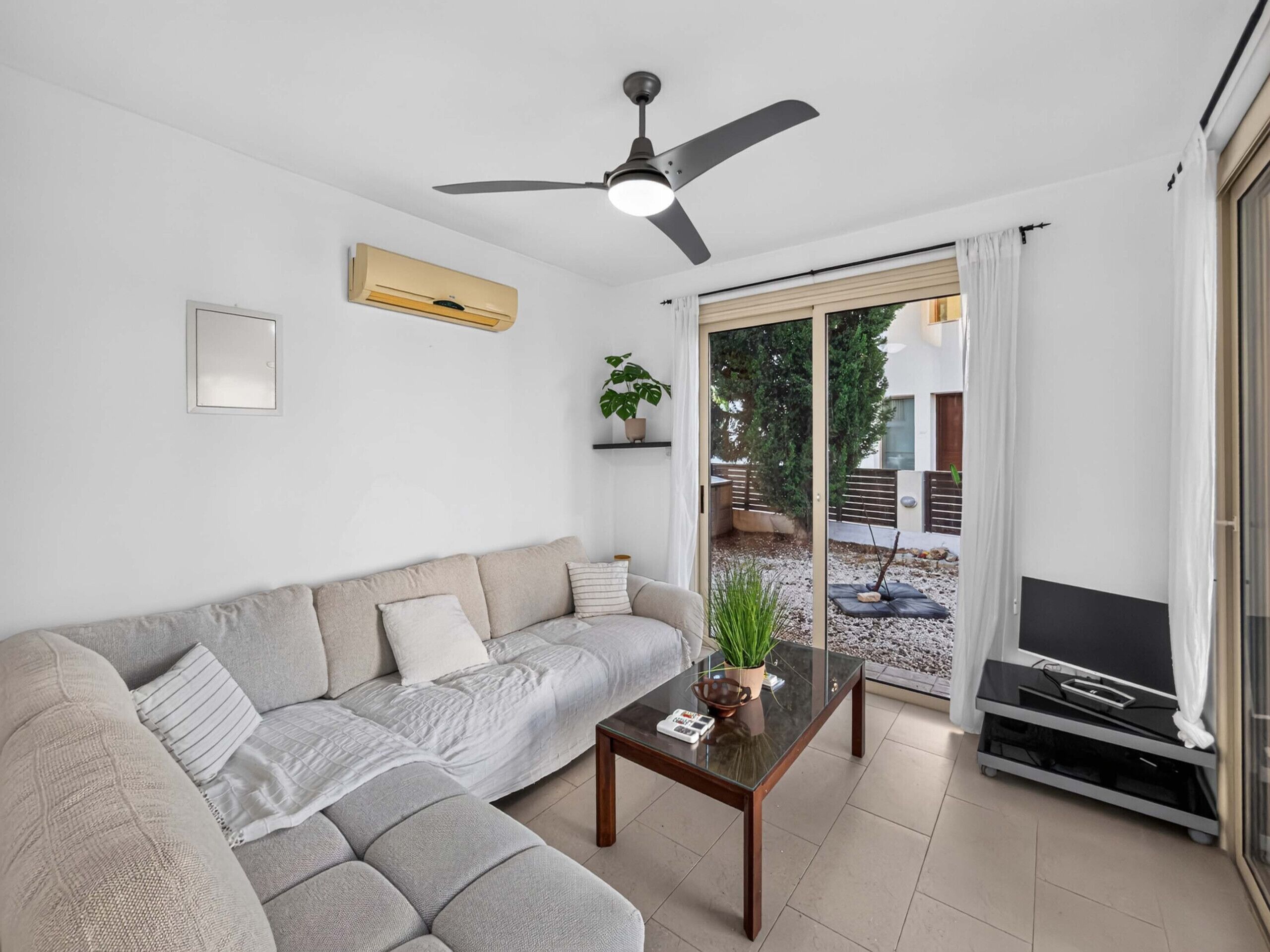 Un salon moderne avec un canapé d’angle, une table basse en verre, un ventilateur de plafond et une porte-fenêtre coulissante donnant sur un espace extérieur.