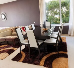 A modern living room with a dining area, featuring a sofa, a dining table with chairs, a decorative mirror, and a view through a large window. - Luxury Seafront Villa | Private Beach, Pool&Garden (Oroklini)