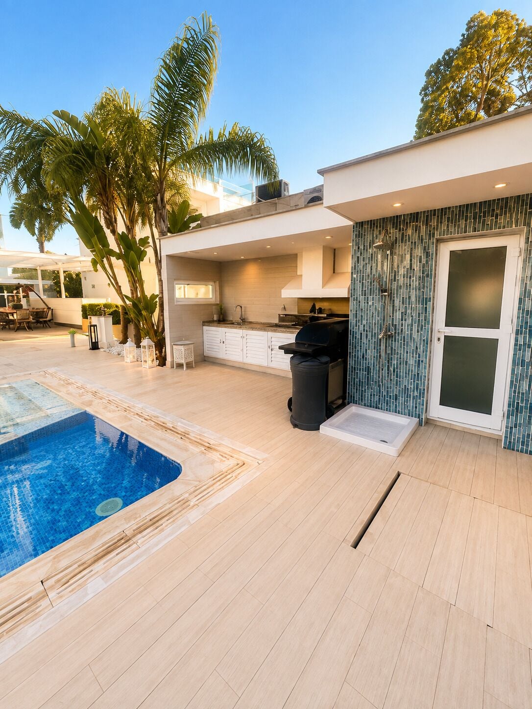 A modern outdoor kitchen with a pool, surrounded by palm trees and a clear sky.