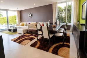 A modern living room with a sofa set, dining table, and chairs, a decorative mirror on the wall, and a view through large windows. - Luxury Seafront Villa | Private Beach, Pool&Garden (Oroklini)