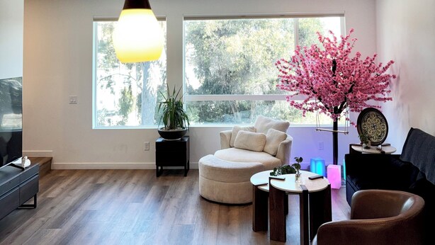 Uma sala de estar moderna com sofá, puf e mesa de centro. Há uma janela grande com vista para árvores, um lustre e um vaso de planta.