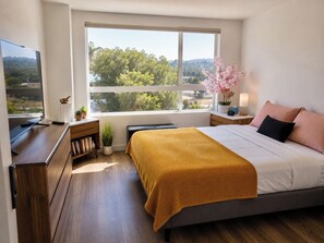 A bedroom with a large window, a bed with a yellow blanket, a bedside table with a lamp, and a TV on a desk. - New House W/ Rooftop Near DTLA, Dodgers, Universal, Griffith Park. Great price! (Los Angeles)