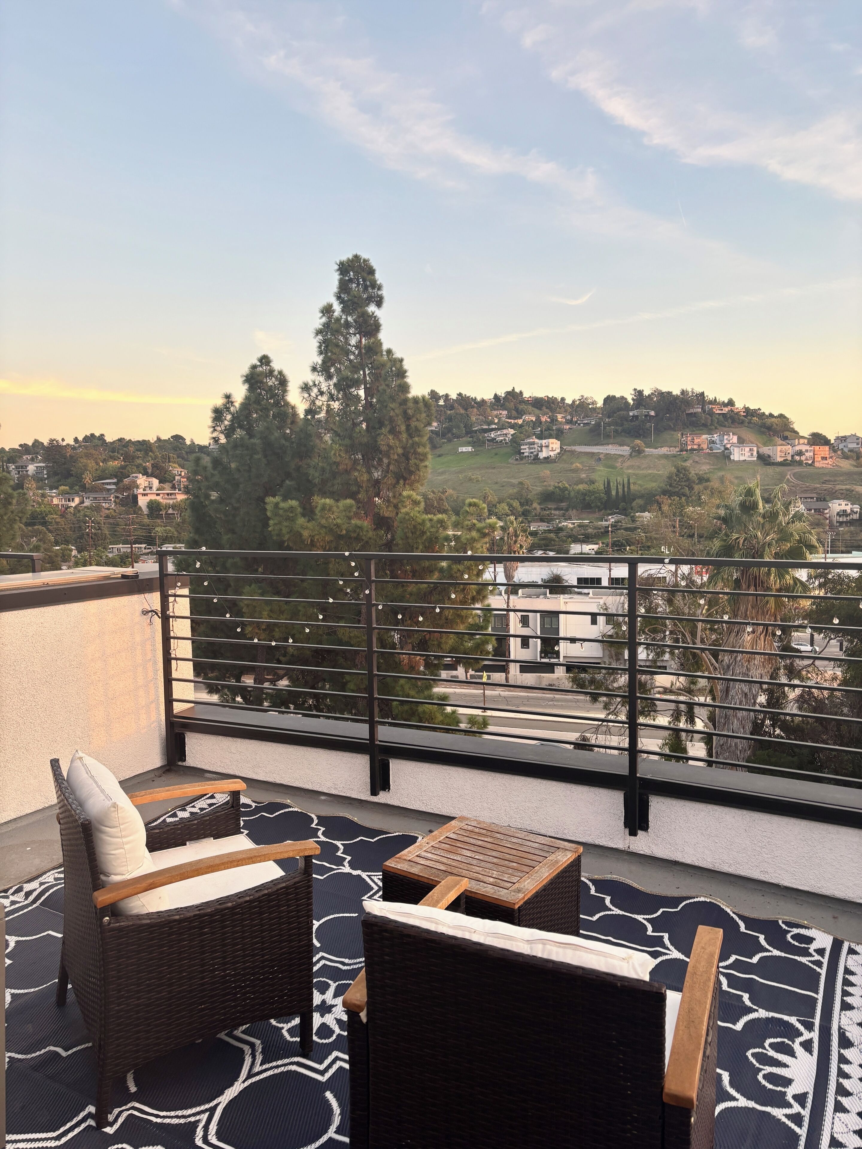 A balcony with wicker furniture, a patterned rug, and a view of a hillside town.