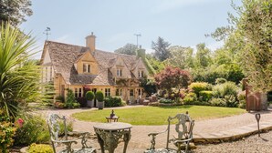 A traditional house with a stone facade, a tiled roof, and a well-maintained garden with a lawn, shrubs, and a patio area with metal chairs and a table. - The Old Forge - Stanton, Broadway (Stanton, nr Broadway)
