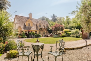 Ein traditionelles Haus mit einer Steinfassade, einem Ziegeldach und einem gepflegten Garten mit Rasenflächen, Sträuchern und einer Terrasse mit Metallstühlen und einem Tisch.