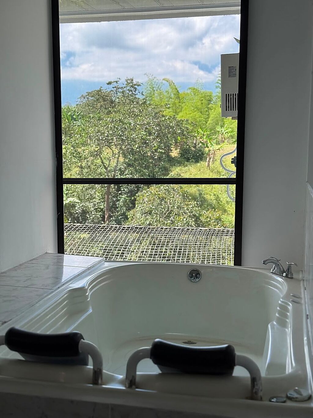 A bathroom with a bathtub, a window with a view of greenery and a road, and a water heater.