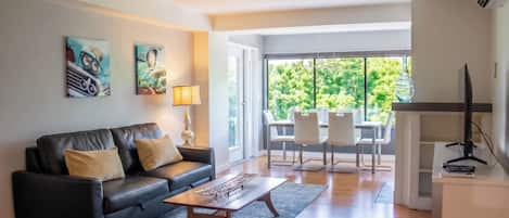 A modern living room with a black leather sofa, a coffee table, and a dining area with white chairs. There is a large window providing a view of greenery outside.