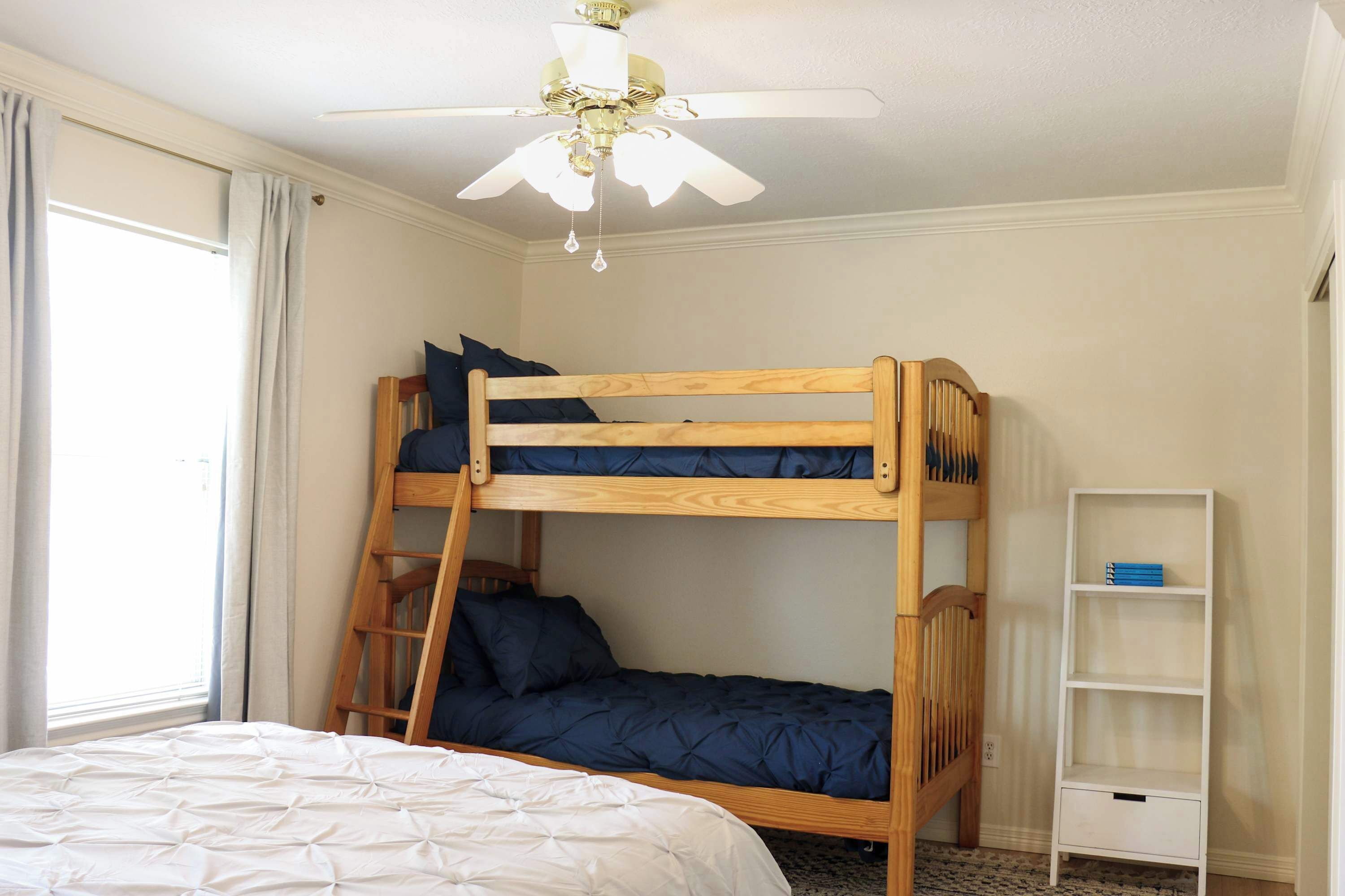 Habitación con literas, ventilador de techo, ventana con cortinas y una estantería blanca.