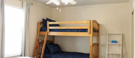Une chambre avec un lit superposé, un ventilateur de plafond, une fenêtre avec des rideaux et une étagère blanche.