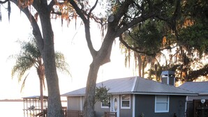 A residential property with a wooden deck, a house with a grey roof, and a body of water in the background. - Cozy cottage in charming St. Cloud perfect for your getaway. (St. Cloud)