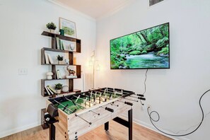 Una habitación con mesa de fútbol de mesa, un televisor de pared que muestra una escena natural y una estantería con diversos objetos.