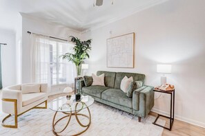 A living room with a green sofa, a white armchair, a glass coffee table, and a potted plant. - Family Retreat 2BD/2BA | Pool |TMC & NRG| Sleeps 8 (Houston)