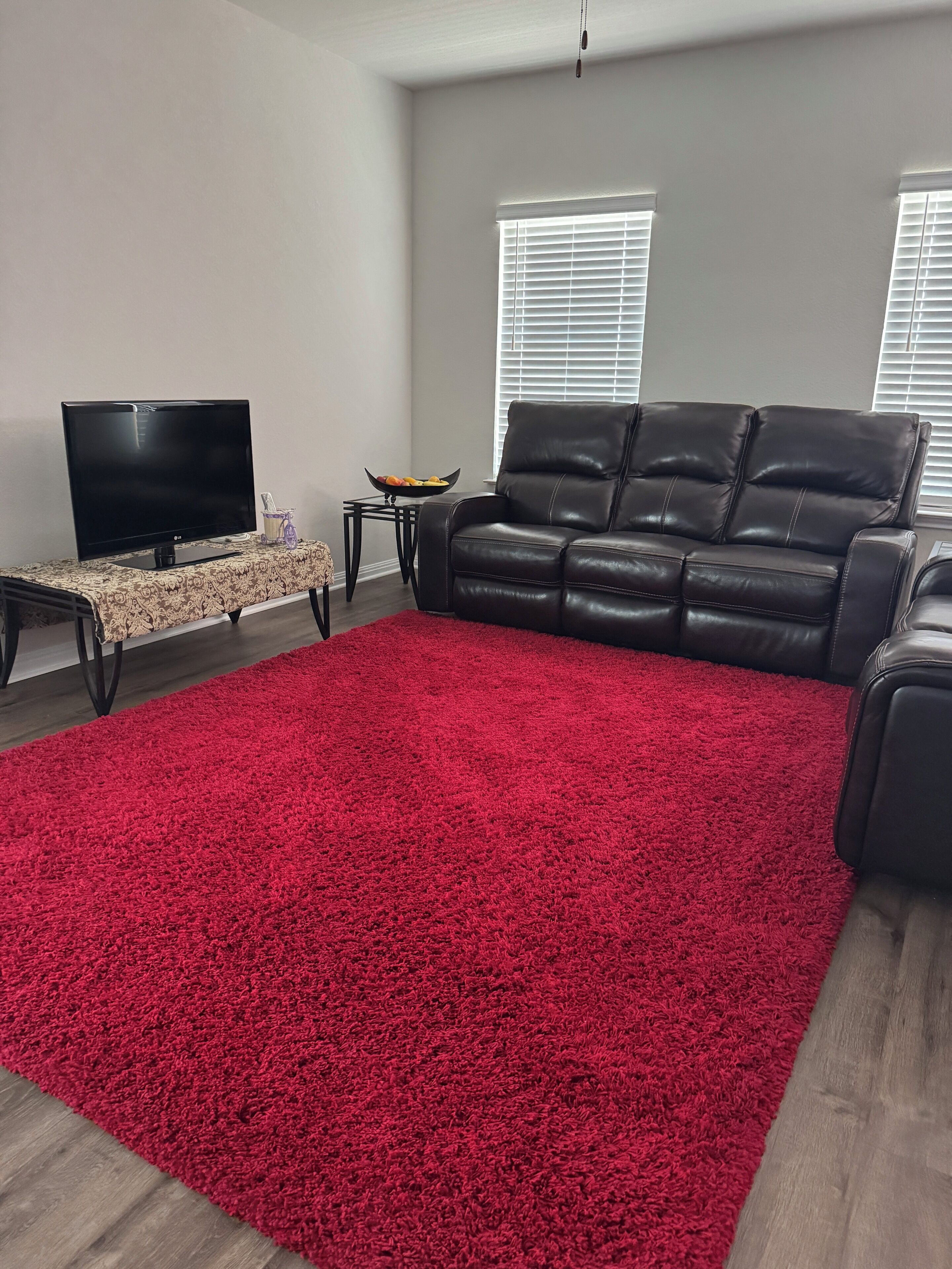 Un salon avec une moquette rouge, un canapé en cuir noir, un téléviseur posé sur un meuble et une petite table sur laquelle se trouve un saladier rempli de fruits.