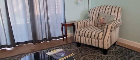 A living room with a striped armchair, a glass coffee table, a patterned carpet, a small wooden side table, and a lamp.
