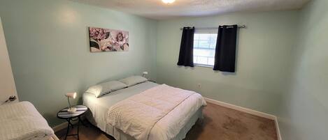 Une chambre à coucher avec un lit, une table de chevet avec une lampe, un tableau accroché au mur et une fenêtre avec des rideaux.