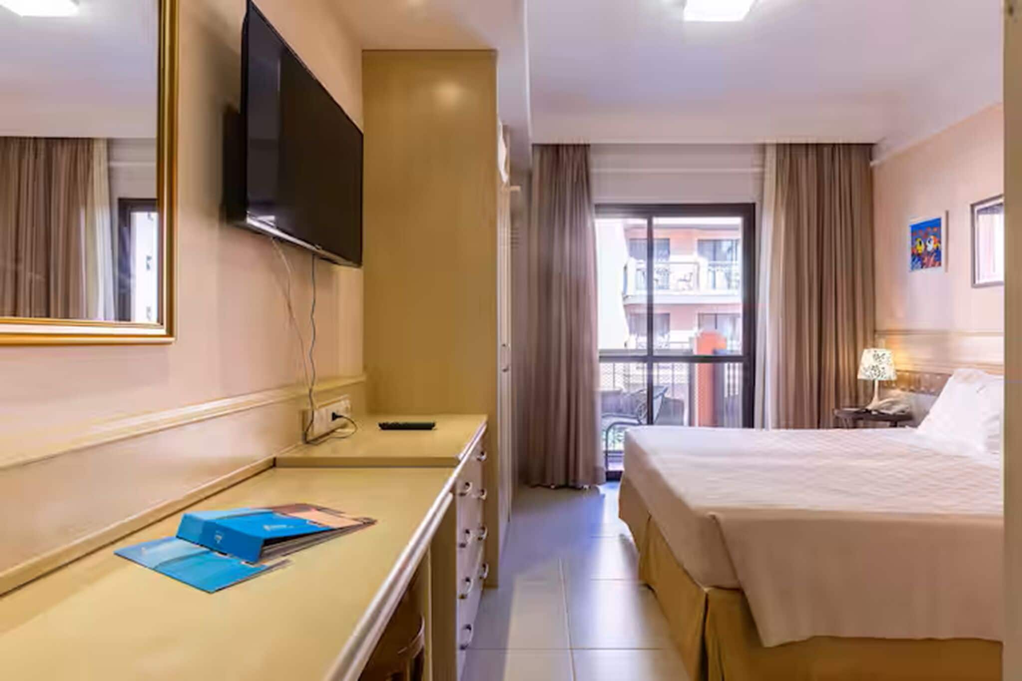 Ein Hotelzimmer mit einem Bett, einem Schreibtisch, einem Flachbildfernseher, einem Spiegel und einem Fenster mit Vorhängen.