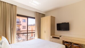 Quarto de hotel com cama, televisão, guarda-roupa de madeira, escrivaninha e varanda com cadeiras.