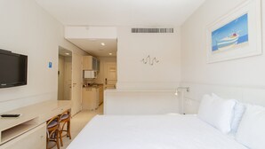 Quarto de hotel com uma cama, uma televisão, uma escrivaninha com cadeiras e uma cozinha compacta ao fundo.