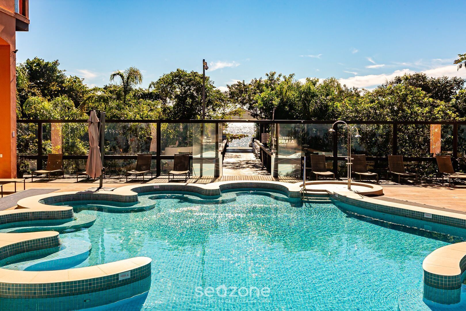A swimming pool with a glass barrier, lounge chairs, and a view of a natural landscape.