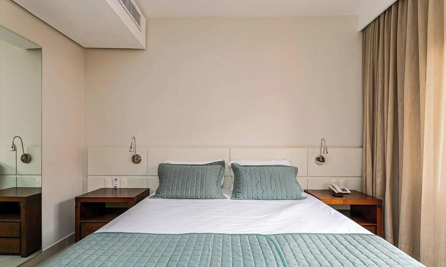 A hotel room with a bed, two bedside tables, a wall-mounted phone, and a window with curtains.