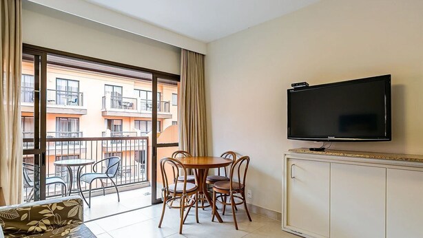A balcony with a table and chairs, a television on a cabinet, and a view of a building with balconies. - Apt W/pool in Luxury Resort in Jurere Jbv167 (Florianópolis)