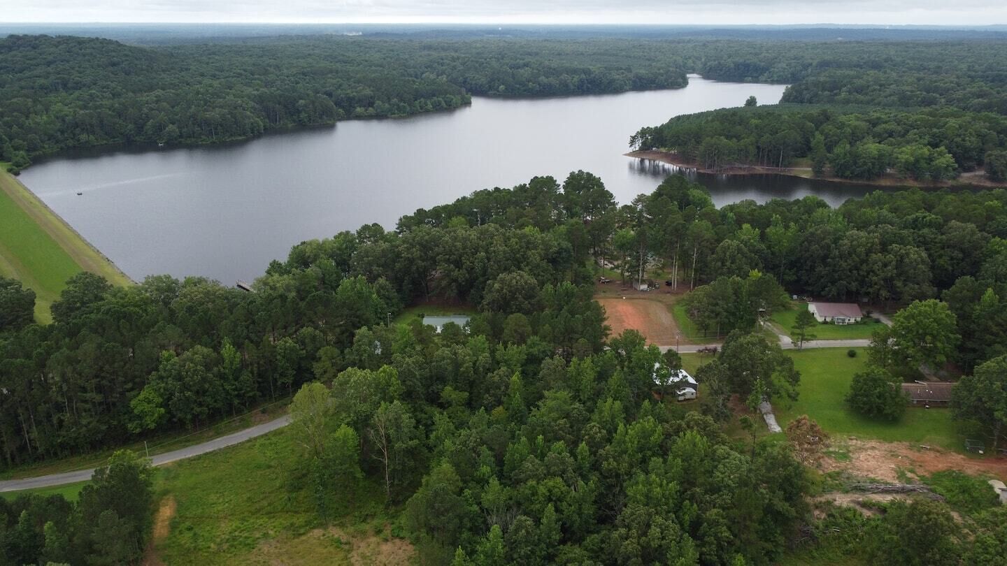 Vista aérea de un lago rodeado de bosque denso, con algunos edificios y una carretera a la vista.