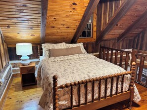 A wooden loft bedroom with a sloped ceiling, a bed with a floral-patterned bedspread, a nightstand with a lamp, and a wooden railing.