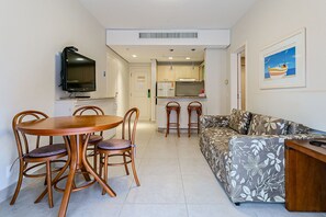 Ein Wohnzimmer mit Sofa, Esstisch mit Stühlen, Fernseher und Küchenzeile mit Kühlschrank und Mikrowelle.