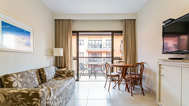 A hotel room with a sofa, a TV, a dining table with chairs, and a balcony with a view of buildings. - Suite w/ Pool View in Luxury Resort Jurere Jbv348 (Florianópolis)