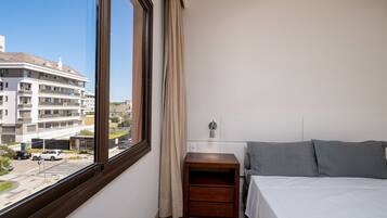 Quarto de hotel com uma janela grande, uma cama, uma mesa de cabeceira e um criado-mudo.