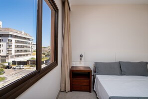 Quarto de hotel com uma janela grande, uma cama, uma mesa de cabeceira e um criado-mudo.