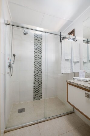 Een douche met glazen wanden, een betegelde wand, een wastafel met een marmeren aanrechtblad en een spiegel met handdoeken.