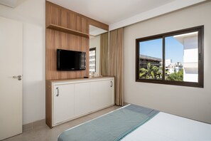 A hotel room with a bed, a TV mounted on a wooden cabinet, a window with a view of buildings, and a door. - Spectacular Suite in a Luxurious Resort Jbv179 (Florianópolis)