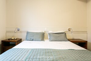 Uma cama com colcha acolchoada, duas almofadas, um telefone ao lado da cama e uma mesinha de cabeceira.