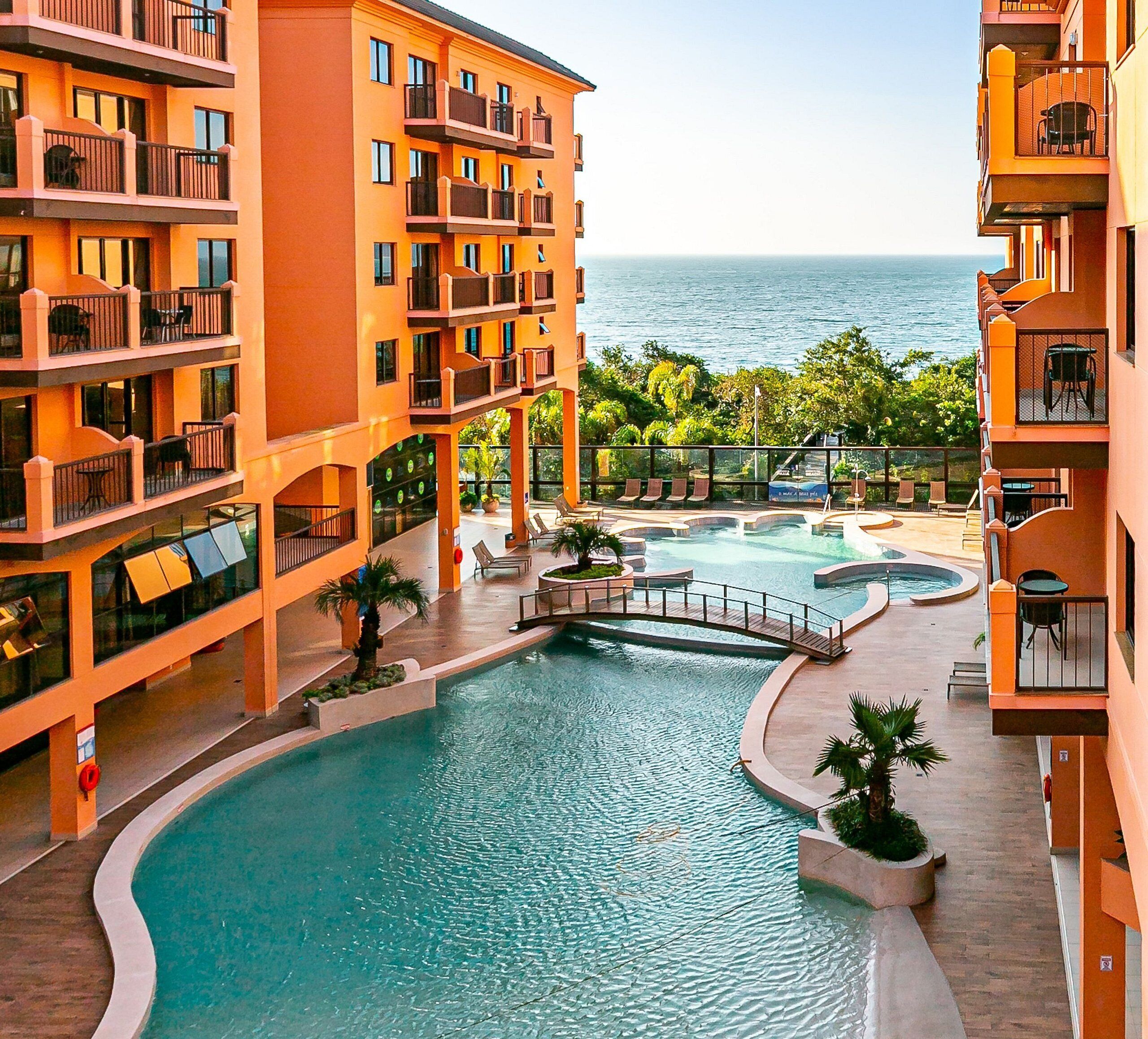 A multi-story hotel with a central swimming pool, outdoor seating, and a view of the ocean.