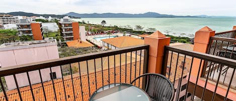 A balcony with a glass table and chairs, overlooking a pool and a body of water with distant mountains.