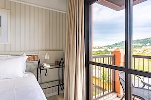 Ein Hotelzimmer mit einem Bett, einem Nachttisch mit Telefon, einer Glasablage und Blick auf einen Balkon mit Gartenmöbeln.