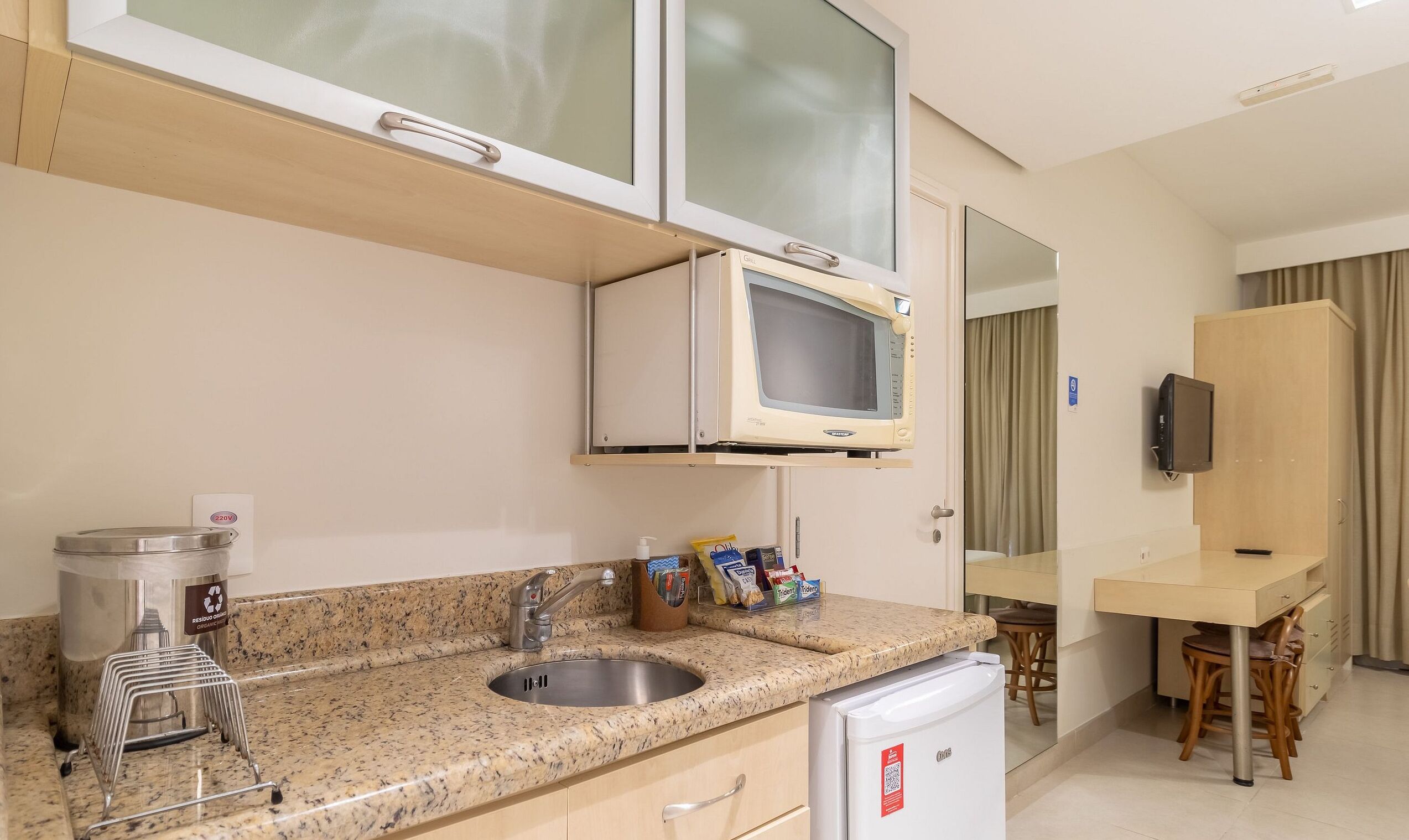Uma cozinha de hotel compacta, com micro-ondas, pia e pequena geladeira.