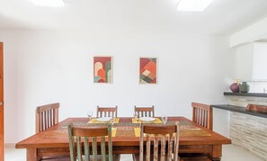 Uma área de refeições com mesa e cadeiras de madeira, duas pinturas abstratas na parede e uma bancada de cozinha com objetos decorativos.