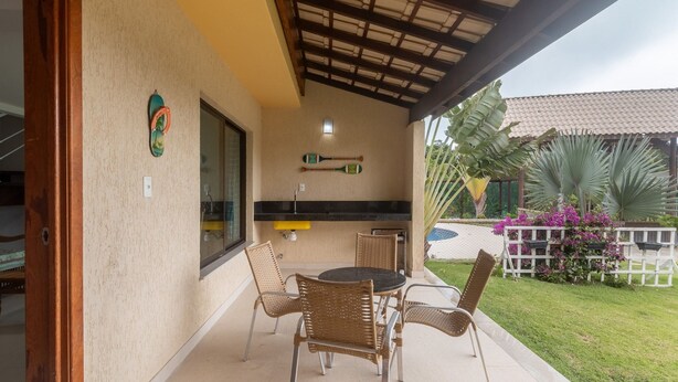 A covered patio with wicker furniture, a small table, and a window with a view of the garden. - House in Cond. W/pool, 500 m From the Sea Atp0001 (Porto Seguro)