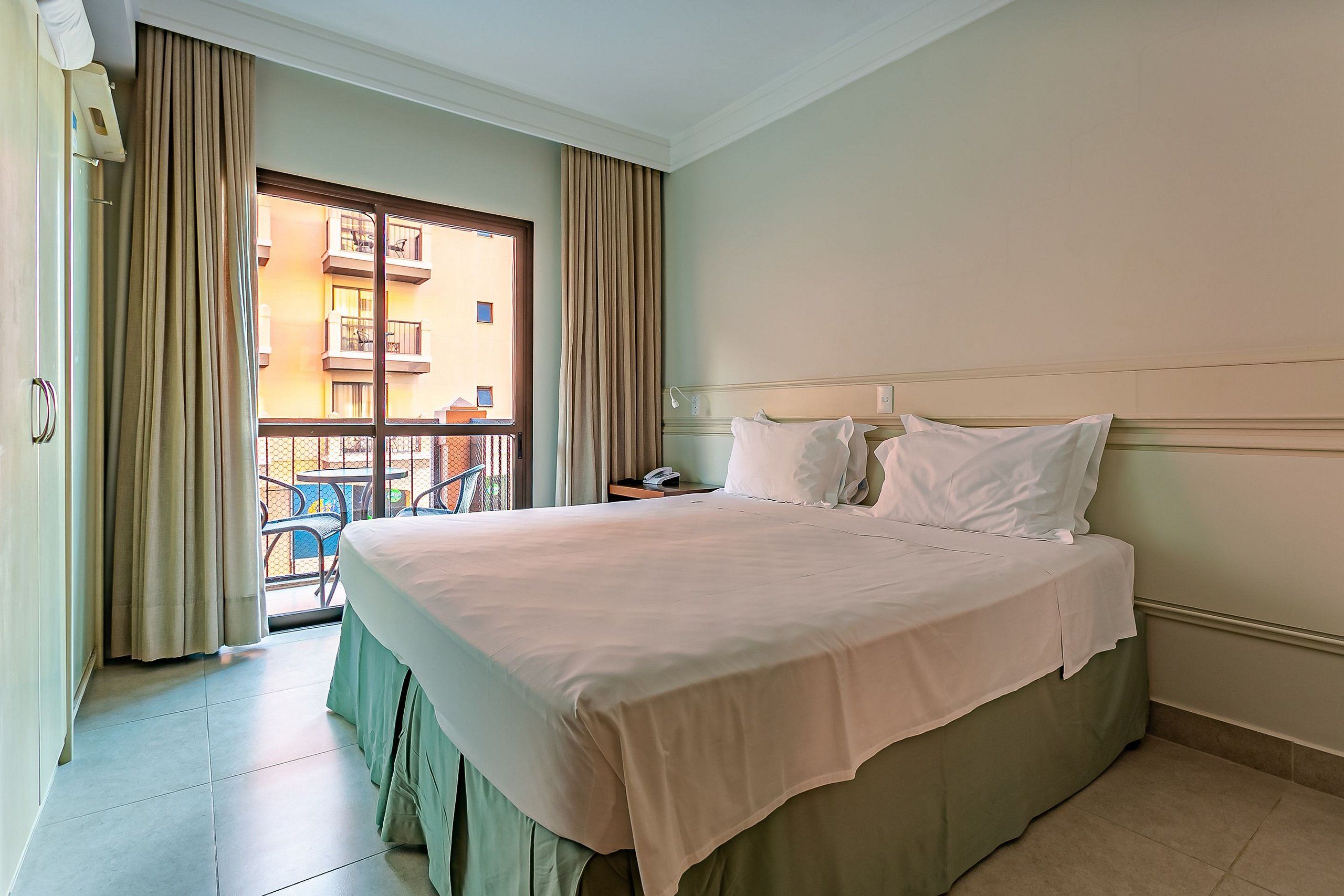 A hotel room with a large bed, a window with curtains, a balcony, and a view of buildings.
