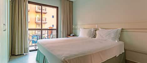 A hotel room with a large bed, a window with curtains, a balcony, and a view of buildings.