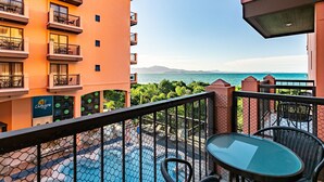 Un balcón con una mesa y sillas de vidrio, con vista a una piscina y al mar y las montañas.