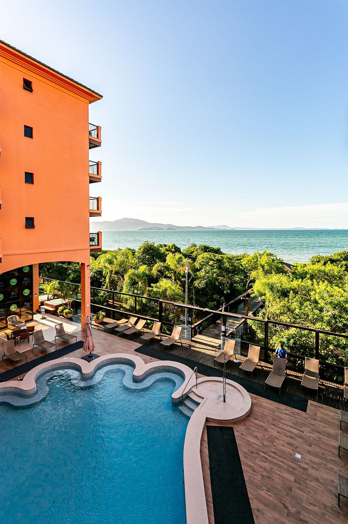 Área de piscina com vista para o mar, cercada por vegetação e um edifício com varandas.