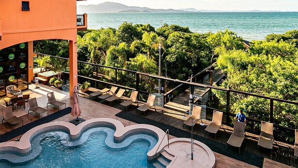 Zona de piscina con vistas al mar, rodeada de vegetación y un edificio con balcones.