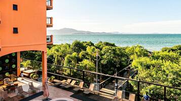Área de piscina com vista para o mar, cercada por vegetação e um edifício com varandas.